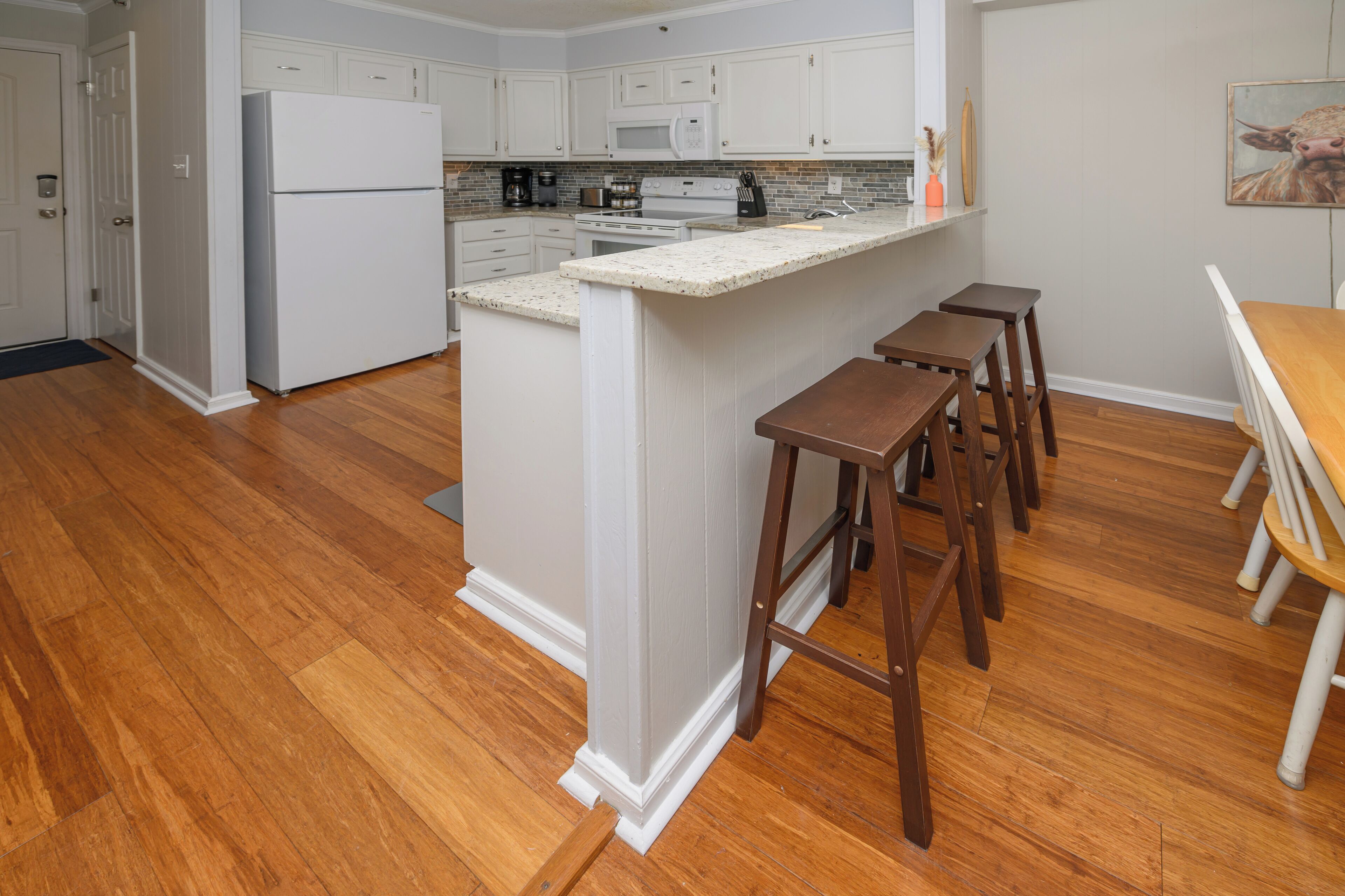 A kitchen with white cabinets, a double oven, a microwave, and a refrigerator. There are wooden stools and a wooden table.