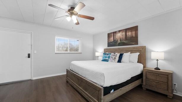 Une chambre avec un lit en bois, un ventilateur de plafond, une fenêtre avec des stores, une table de chevet avec une lampe et un tableau accroché au mur.