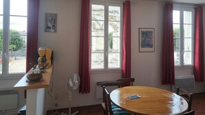 Une pièce avec une table ronde en bois, deux chaises, un ventilateur, une machine à café et une fenêtre avec des rideaux rouges.