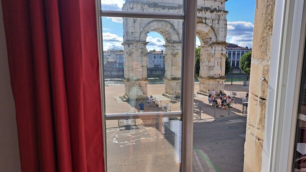 Une vue d’une arche antique, aperçue à travers une fenêtre, avec des personnes assises à l’extérieur.