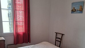 Une chambre à coucher avec un lit, une table de chevet, une lampe, une fenêtre avec des rideaux rouges et un tableau accroché au mur.
