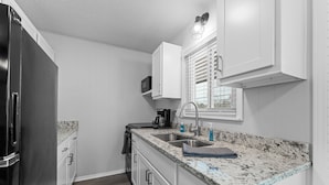 A compact kitchen with white cabinetry, a granite countertop, a stainless steel refrigerator, and a window with blinds.