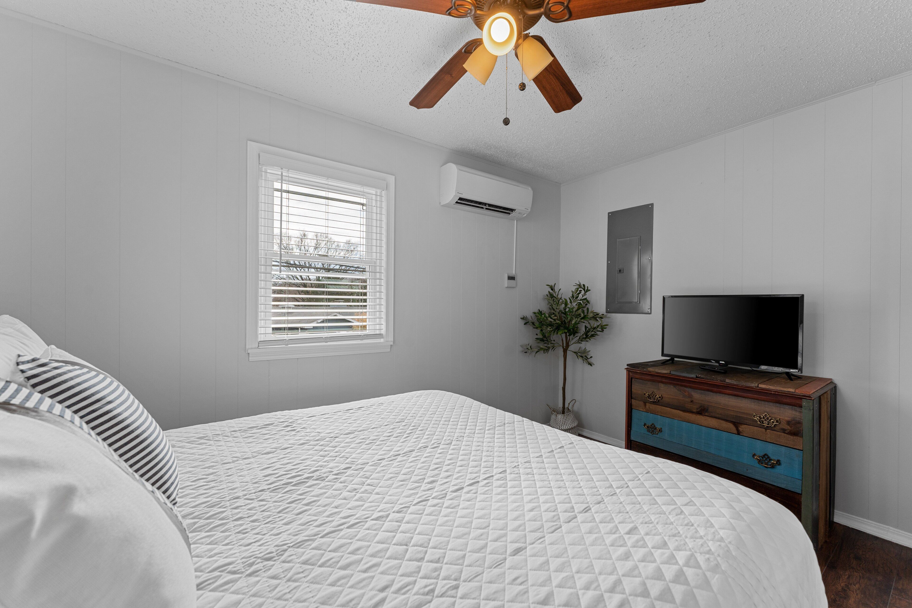 Un dormitorio con cama, ventilador de techo, unidad de aire acondicionado, televisor sobre una cómoda y una ventana con persianas.