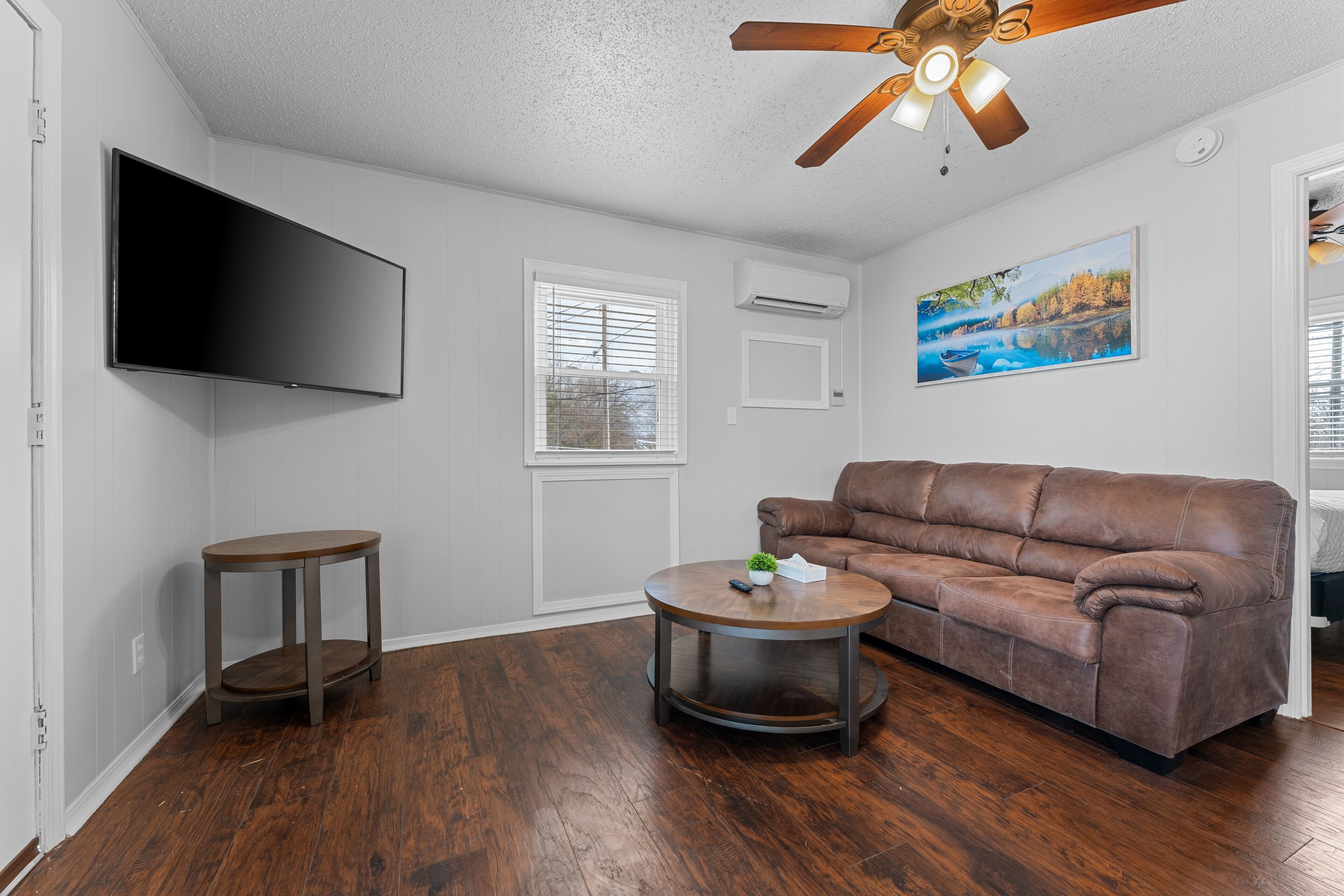 A living room with a brown leather sofa, a round coffee table, a flat-screen TV mounted on the wall, a ceiling fan, and a window with blinds.