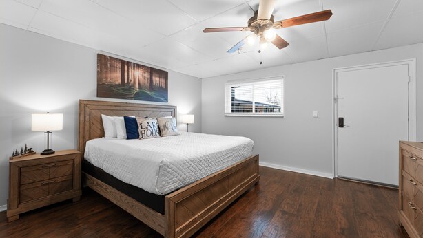 A bedroom with a wooden bed, a nightstand with a lamp, a ceiling fan, a window with blinds, and a door. - Sc8- Silver Cove 8 1 Bedroom Condo (Branson)