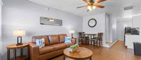 Ein Wohnzimmer mit braunem Ledersofa, einem runden Holztisch, einem Deckenventilator, einem Essbereich mit Tisch und Stühlen sowie einer Küchenzeile mit Mikrowelle.