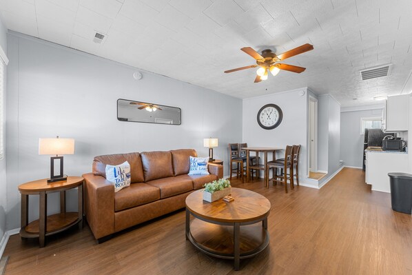 A living room with a brown leather sofa, a round wooden coffee table, a ceiling fan, a dining area with a table and chairs, and a kitchen area with a microwave.