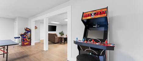 A room with a ping pong table, a basketball arcade game, and a bar area with stools.