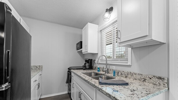 Una cocina compacta con muebles blancos, encimera de granito, refrigerador de acero inoxidable y ventana con persianas.