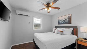 Un dormitorio con cama, ventilador de techo, televisor montado, ventana con persianas y un cuadro en la pared.