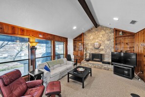 A spacious living room with a stone fireplace, a television, and a large window overlooking a scenic view. - Lake House At Granbury 3 Bedroom Home (Granbury)