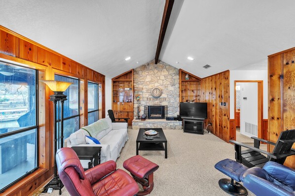 A spacious living room with a stone fireplace, wooden walls, and large windows offering a view. - Lake House At Granbury 3 Bedroom Home (Granbury)