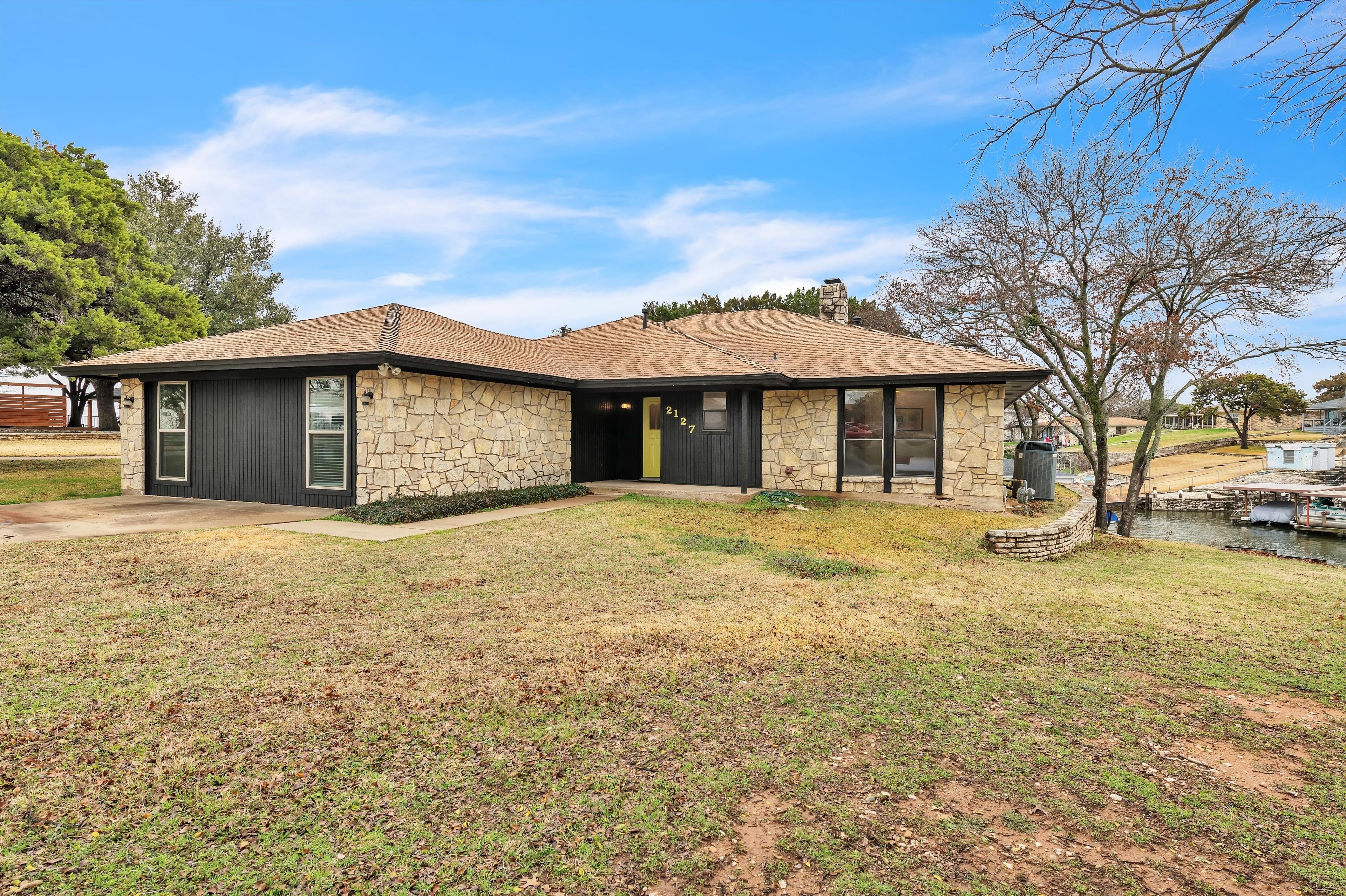 A single-story house with a stone facade and a dark roof, located near a body of water.