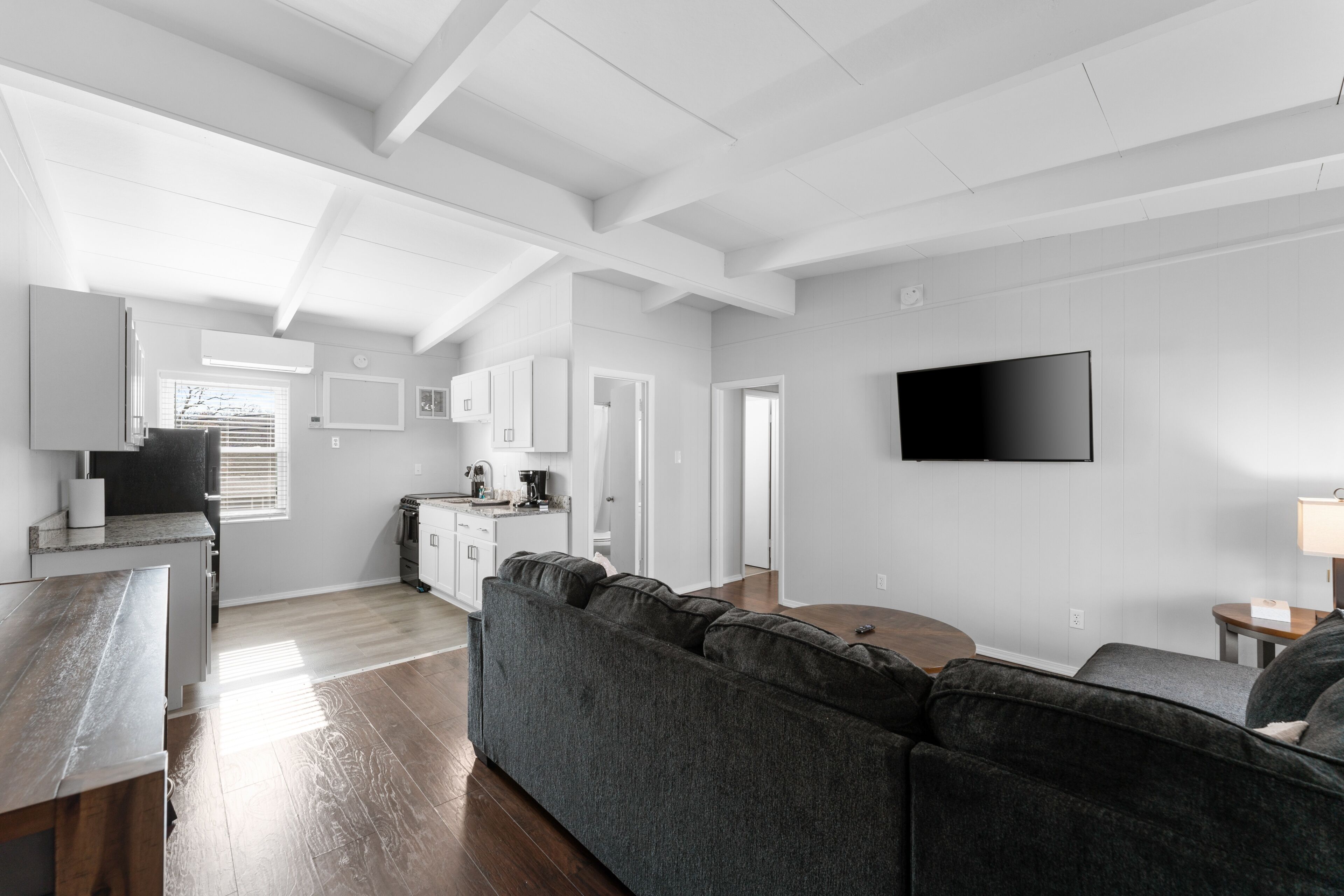 A modern living room with a sectional sofa, a flat-screen TV mounted on the wall, and a kitchen area with white cabinetry.