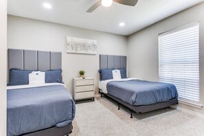 A bedroom with two beds, a nightstand, a ceiling fan, and a window with blinds. - Texas - Reston 1513 5 Bedroom Home (Richardson)