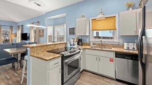 A modern kitchen with a granite countertop, stainless steel appliances, and a dining area with a blue sofa. - Serenity Now-Free Golf Cart-Hot Tub-Comm Pool (Destin)