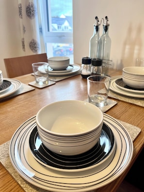 A dining table set with stacked plates, bowls, and glasses, with salt and pepper shakers and a bottle of water. - Natterjack Cottage - Access to the Western Lakes (Millom)