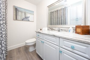 A bathroom with a white toilet, a marble sink, and a large mirror. There is a framed picture on the wall and a patterned shower curtain. - Phoenix On The Bay 1513 3 Bedroom Condo (Orange Beach)