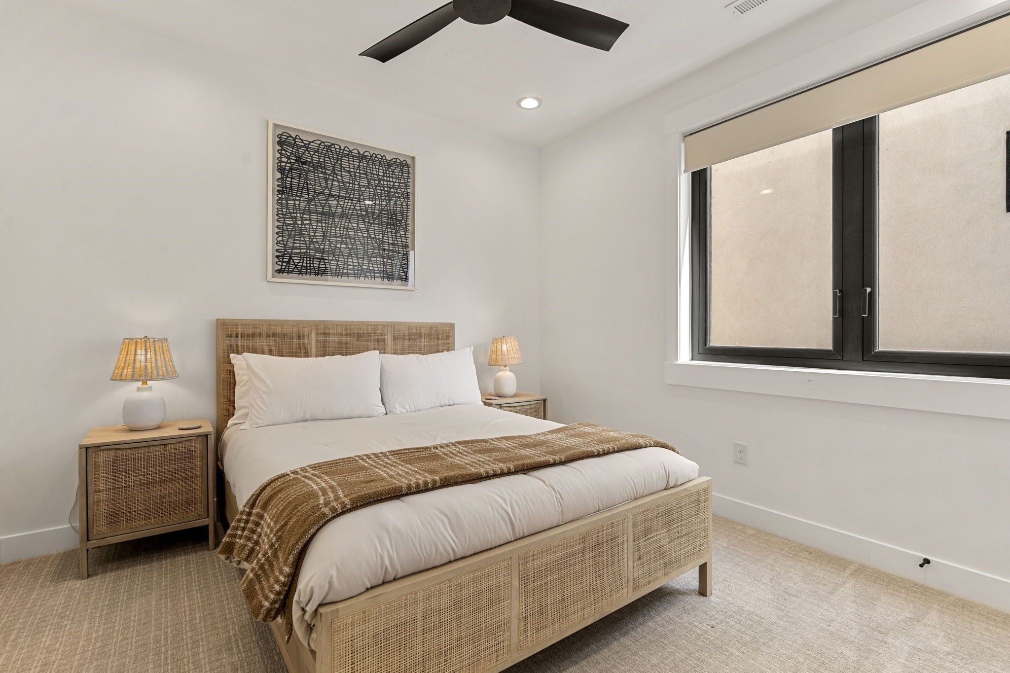 A bedroom with a large bed, two bedside tables with lamps, a ceiling fan, a window with blinds, and a piece of abstract art on the wall.