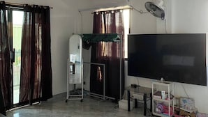 A living room with a flat-screen TV, a ceiling fan, a standing fan, a TV stand with a remote, and a window with curtains.