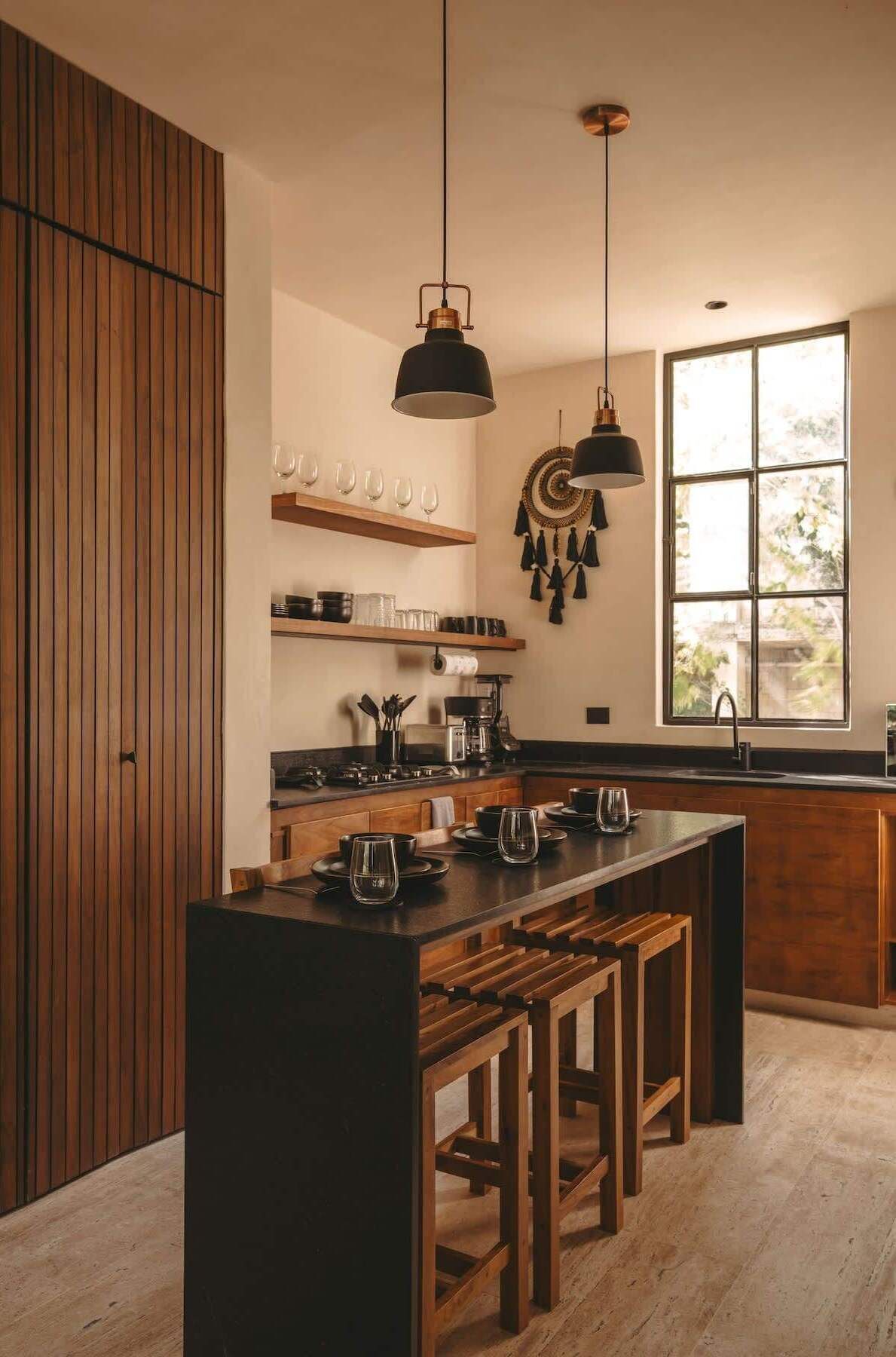 Una cocina moderna con armarios de madera, una isla central con taburetes, iluminación colgante y un amplio ventanal que deja entrar luz natural.
