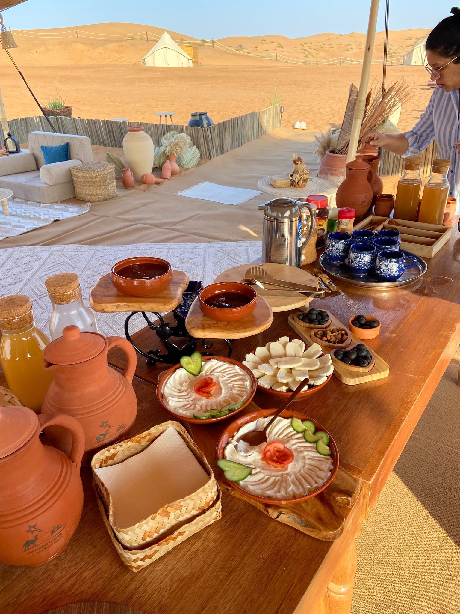 Un campamento en el desierto con una mesa de comedor dispuesta con diversos platos, incluyendo salsas y pan, y una vista de tiendas de campaña y dunas de arena al fondo.