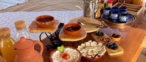 Un campamento en el desierto con una mesa de comedor dispuesta con diversos platos, incluyendo salsas y pan, y una vista de tiendas de campaña y dunas de arena al fondo.