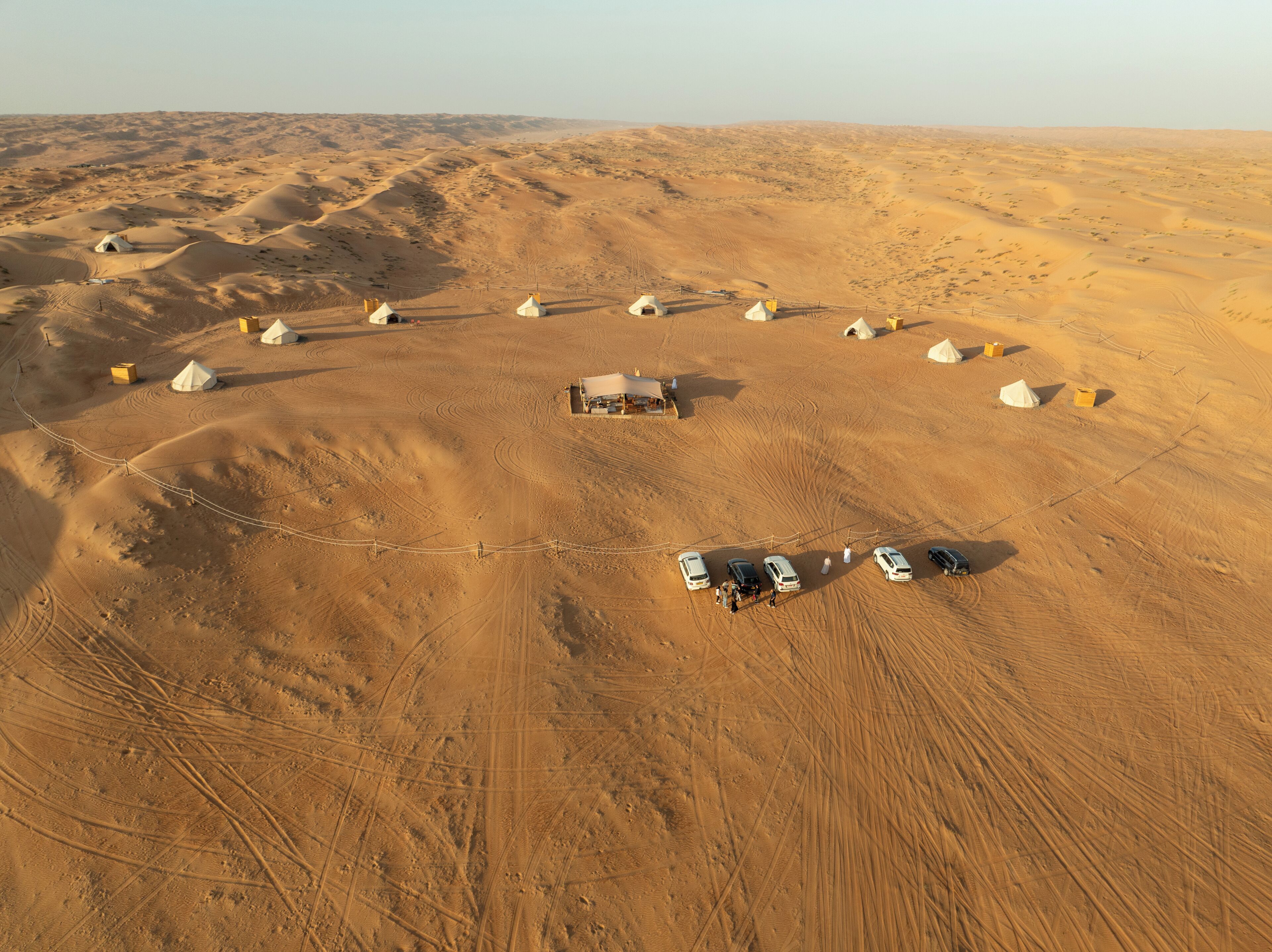 Vista aérea de un campamento en el desierto con yurtas y un edificio central, rodeado de vehículos y una zona vallada.