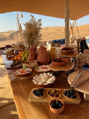 A desert camp with a dining table set with food and drinks, surrounded by tents and a view of sand dunes. - Twilight dunes By Miracle Oman (Bidiya)