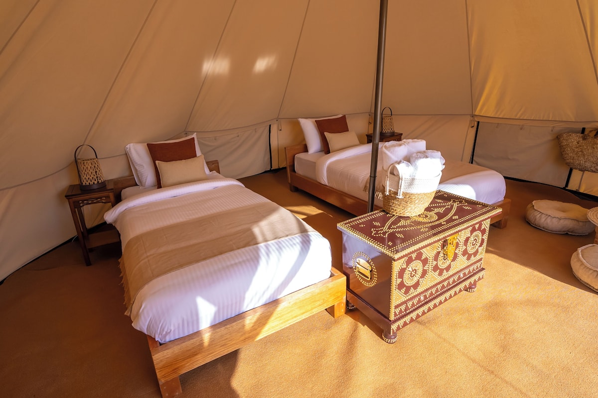 A tent with two beds, each with white bedding and wooden frames. There is a small table with a woven basket in the center.
