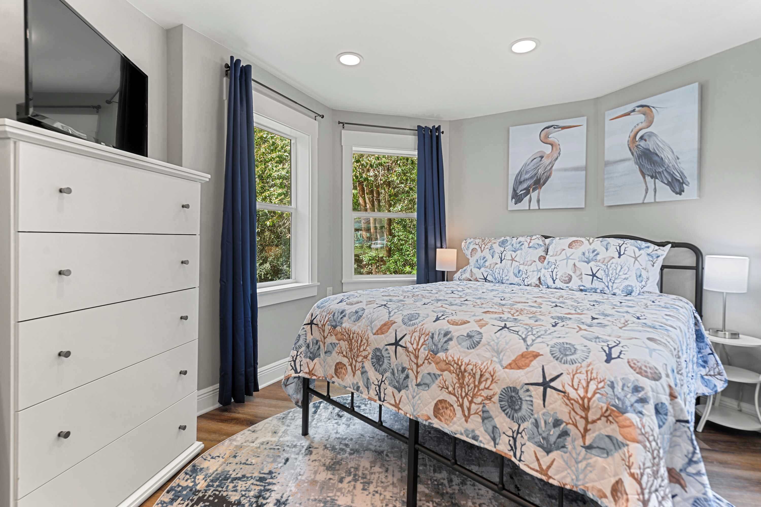 Een slaapkamer met een bed, een dressoir, een raam met gordijnen en twee aan de muur hangende schilderijen van vogels.