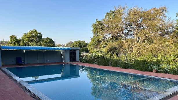 Una piscina rettangolare con superficie blu e trasparente, circondata da un solarium piastrellato di colore rosso. È presente un'area relax coperta con tetto blu e un ingresso che conduce a uno spazio chiuso.