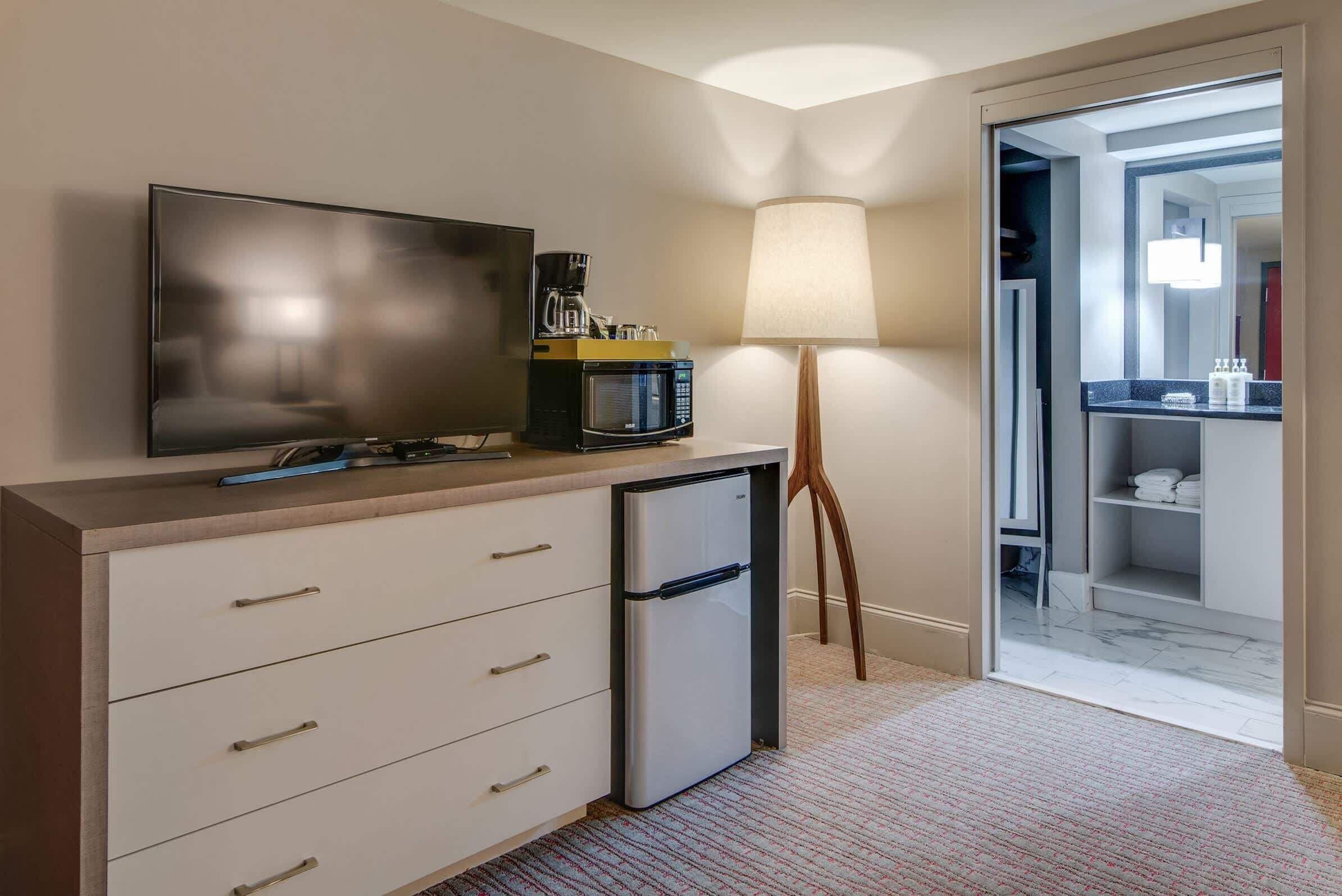 Une chambre d’hôtel compacte équipée d’une télévision à écran plat, d’une machine à café, d’un mini-réfrigérateur, d’une lampe et d’un dressing.
