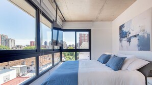 Un dormitorio con un ventanal grande, una cama con ropa de cama blanca y azul, y vista a la ciudad.