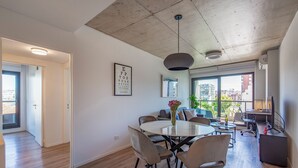 Una sala de estar moderna con una mesa de comedor redonda, asientos cómodos, un ventanal con vistas a la ciudad y un techo con concreto expuesto.