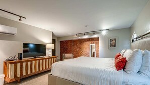 A modern hotel room with a large bed, a TV on a wooden console, a brick accent wall, and a ceiling light fixture. - Charming Stay for 4 in DT, Walk to Broadway! (Nashville)