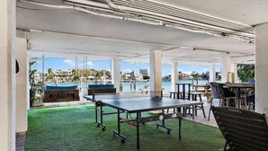 An outdoor seating area with tables and chairs, a ping pong table, and a view of the water and buildings in the background.