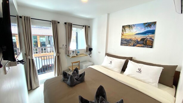 A hotel room with a large bed, a flat-screen TV mounted on the wall, a small table with a lamp, and a window with curtains. - SJ Beach House Patong (Patong)