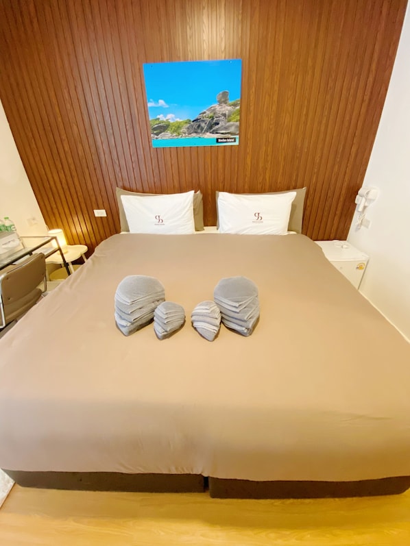 A hotel room with a wooden headboard, a bed with a beige bedspread, two pillows, and a stack of folded towels. - SJ Beach House Patong (Patong)