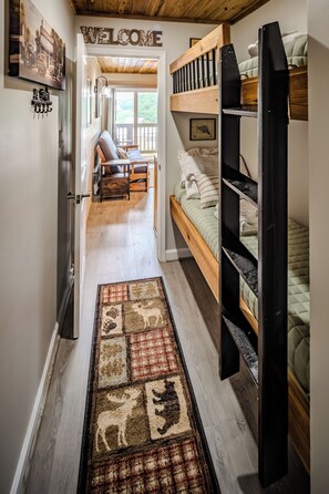 A narrow hallway with a wooden floor, a welcome sign, a bunk bed, a sofa, and a rug with animal prints. - Sugar Ski Comfort One Bedroom - Unit 123 (Sugar Mountain)