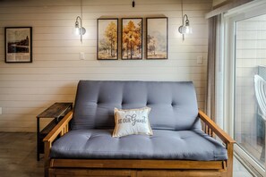 A grey sofa with a cushion that reads 'BE OUR GUEST' in a hotel room with wooden walls, a framed picture of a bridge, and two wall-mounted light fixtures. - Sugar Ski Comfort One Bedroom - Unit 123 (Sugar Mountain)