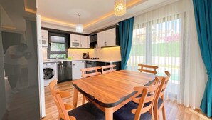 A compact kitchen with modern appliances, a dining area with a wooden table and chairs, and a view of a garden through large windows.