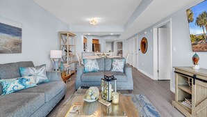 Een moderne woonkamer met een grijze bank, een salontafel met decoratieve objecten, een boekenkast en een schilderij van een strandlandschap.