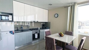 A modern kitchen with white cabinets, a microwave, stove, and dishwasher. There is a dining table with chairs and a fruit bowl. A clock is on the wall. - Flat in Praia Plateau (Praia)
