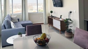 A modern living room with a sofa, a dining table with a fruit bowl, a TV mounted on the wall, and large windows with curtains. - Flat in Praia Plateau (Praia)