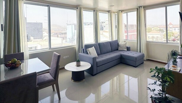 A modern living room with a grey sectional sofa, a dining table with chairs, and a small round table with a plant. Large windows offer a view of the cityscape. - Flat in Praia Plateau (Praia)