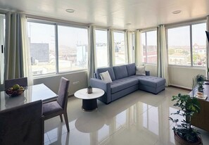 A modern living room with a grey sectional sofa, a dining table with chairs, and a small round table with a plant. Large windows offer a view of the cityscape. - Flat in Praia Plateau (Praia)