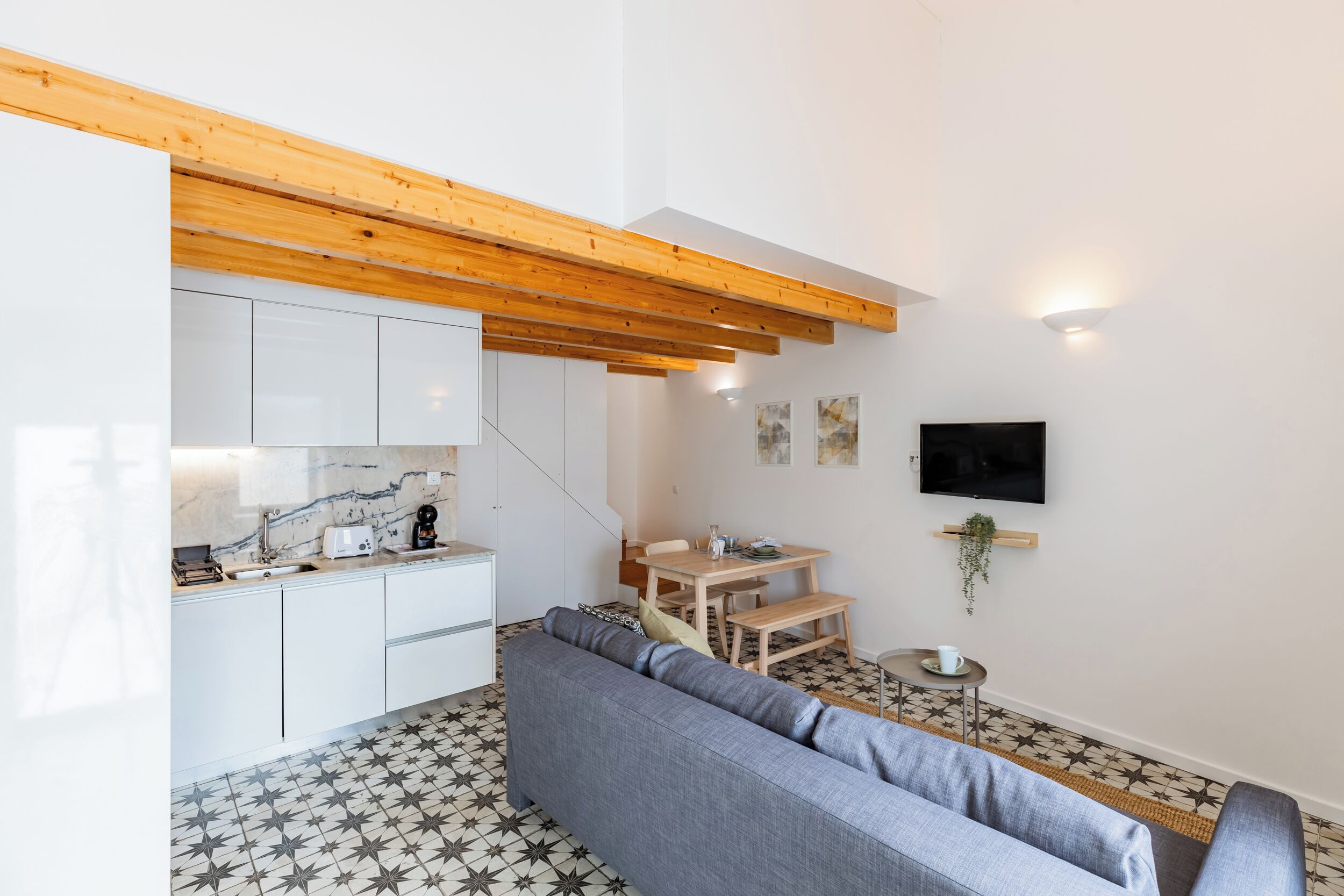 Una cucina moderna con mobili bianchi, soffitto in legno e pavimento piastrellato. Si vede un divano grigio e un tavolino con una pianta.