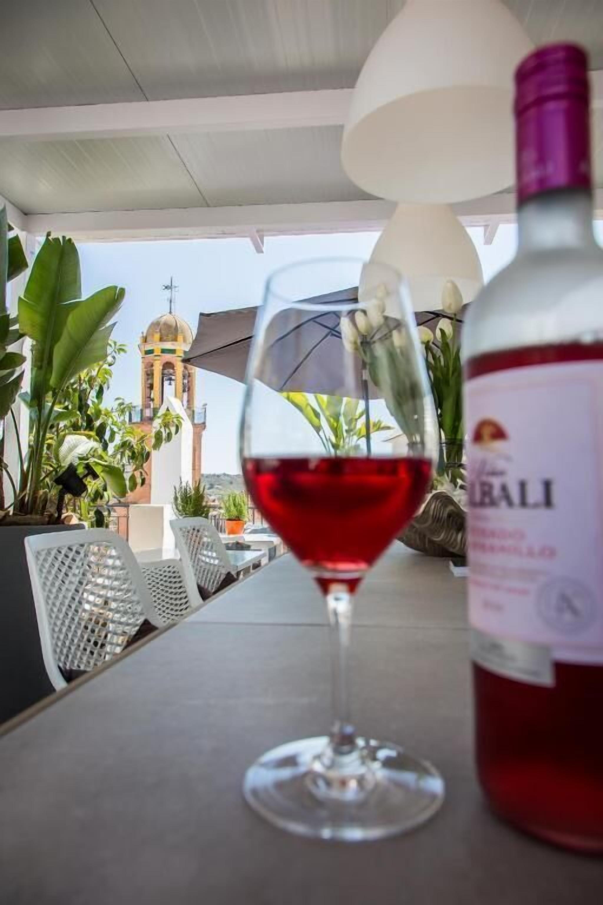 Una mesa con una copa de vino y una botella, rodeada de plantas en macetas y mobiliario de exterior.