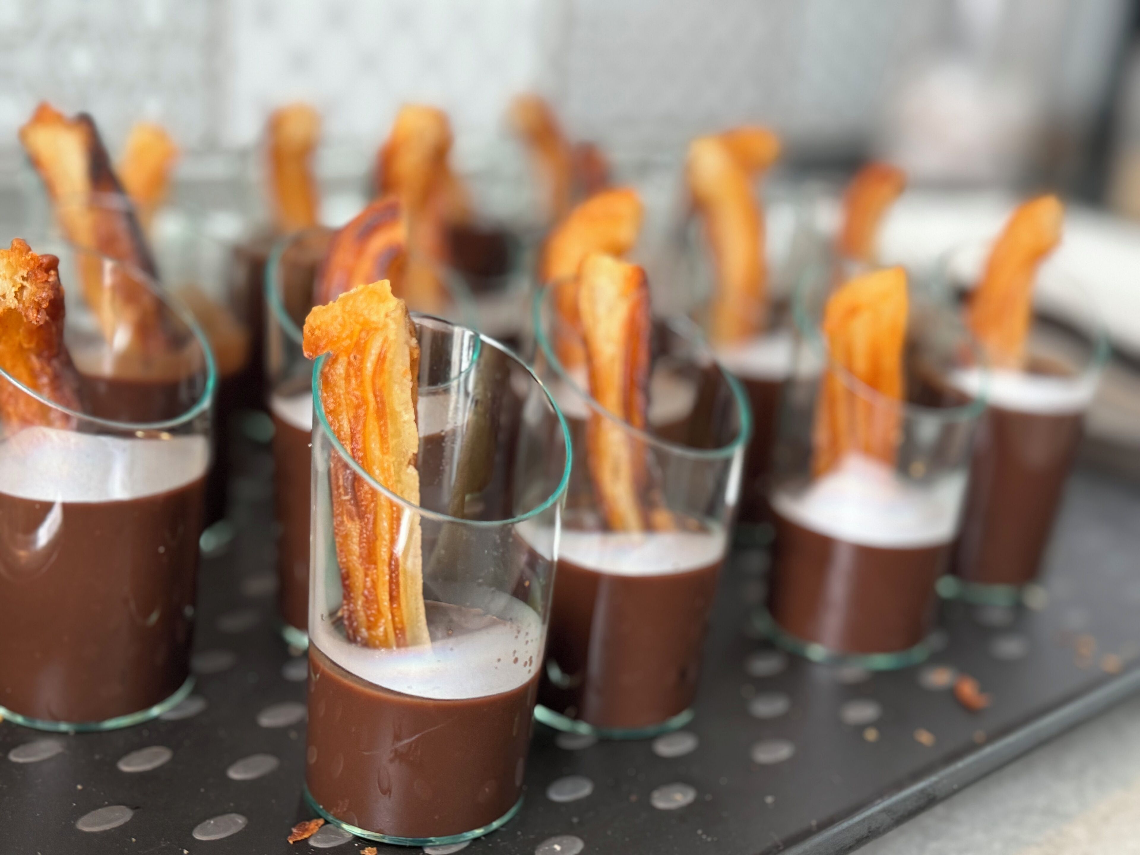 Varios vasos transparentes llenos de un líquido oscuro, cada uno coronado con una porción de tocino crujiente.